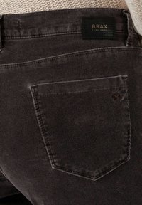 Poche arrière d'un pantalon en velours côtelé marron foncé avec étiquette de marque BRAX noire et haut en maille crème visible à la taille.