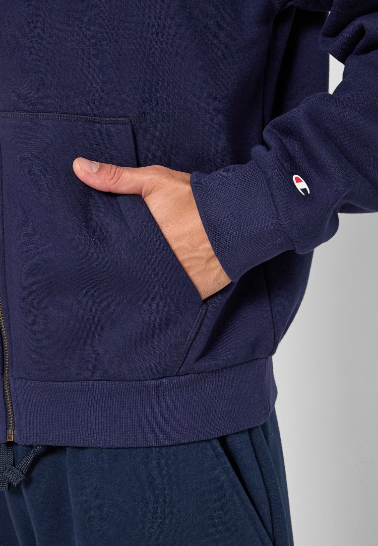 Felpa zip-up blu navy con tasca frontale, polsini e fascia in costina. Presenta una patch con il logo sulla manica. Tessuto morbido e strutturato.