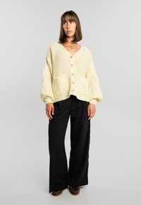 Mujer joven con cabello castaño liso, llevando un cárdigan de punto grueso color crema, pantalones anchos negros, zapatos marrones y collares dorados sobre un fondo gris.