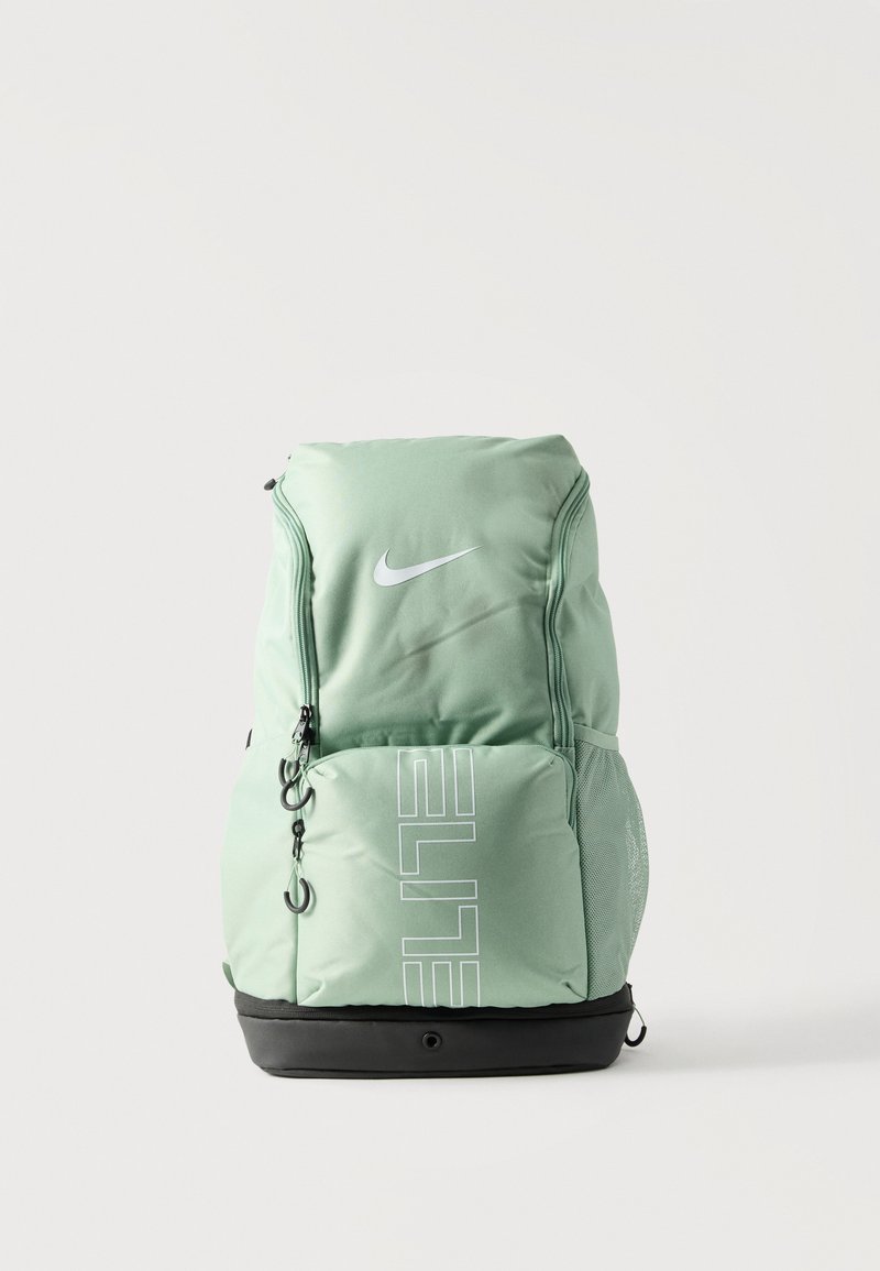 Mochila Nike Elite de color verde claro con múltiples compartimentos con cremallera y una base negra reforzada, mostrada de pie sobre un fondo blanco.