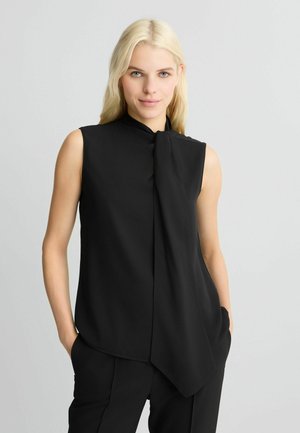 Black sleeveless top with a high neckline and draped tie detail at the front; smooth texture and relaxed fit. Worn with black trousers.