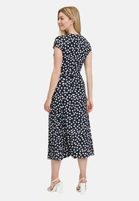 Marinblå klänning med vita prickar, korta ärmar, figurnära bodice och svängande midi-kjol. Modellen bär vita högklackade sandaler.