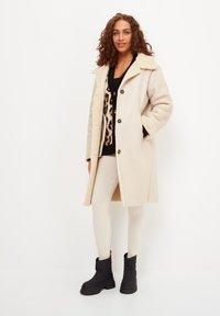 Abrigo de lana beige con un suave cuello, cierre de botones y bolsillos laterales. Lleva puesto un suéter negro con estampado de leopardo y leggings blancos de canalé.