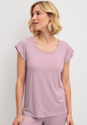Blonde Frau, die ein hellviolettes kurzärmeliges Oberteil und passende Hosen trägt und seitlich vor einem einfarbigen Hintergrund schaut.