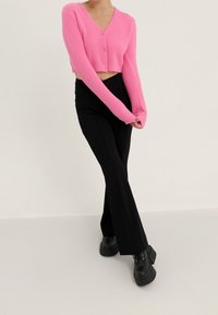 Różowy krótki kardigan z dekoltem w serek i zapięciem na guziki, zestawiony z czarnymi rozszerzanymi spodniami i masywnymi czarnymi butami na platformie. Gładka tekstura na całej powierzchni.