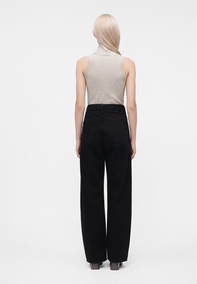 Personne aux longs cheveux blonds portant un haut rayé sans manches et un pantalon noir taille haute à jambes larges, debout de dos sur un fond uni.