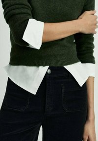 Maglione verde scuro e strutturato indossato sopra una camicia bianca con colletto, abbinato a pantaloni in velluto nero. Bottoni e orli visibili.