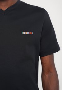 Mustaga lühikese varrukaga T-särk V-kaelusega, millel on vasakul rinnal mitmevärviline tikandiga triip. Sile tekstuur.