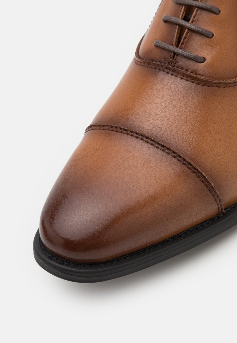 Chaussure habillée en cuir marron avec un bout arrondi, des coutures décoratives et une semelle en caoutchouc noire. Texture lisse avec un léger brillant.