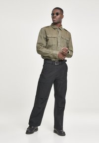 Cămașă cargo de culoare verde măslinie cu două buzunare pe piept, asociată cu pantaloni cargo negri prevăzuți cu buzunare laterale. Include ochelari de soare rotunzi întunecați și pantofi negri.