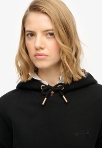 Czarna bluza z kapturem z złotym sznurkiem, miękka w dotyku, z subtelnym detalem logo. Kaptur stylizowany jest na białą koszulę z kołnierzykiem.