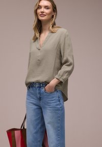 Frau trägt eine hellolivfarbene, strukturierte Bluse und blaue Jeans, hält eine gestreifte rote und pinkfarbene Tragetasche vor einfachem Hintergrund.