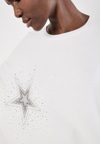 Biała sweter z dzianiny z wyraźnym srebrnym motywem gwiazdy, ozdobionym cyrkoniami oraz mniejszymi, błyszczącymi detalami. Okrągły dekolt.