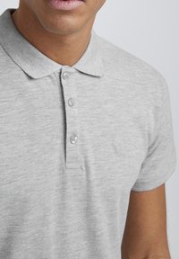Grå polo skjorte laget av teksturert stoff, med krage, tre knapper, og en liten brodert logo på venstre brystområde.
