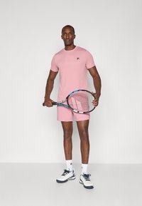 Rosa trenings t-skjorte med logo, matchende shorts, holder en tennisracket, iført hvite og marineblå sko, står på en grå bakgrunn.