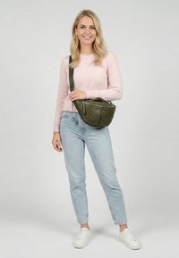Lachende vrouw staat met een grote olijfgroene crossbodytas, draagt een lichtroze trui, blauwe jeans en witte sneakers tegen een neutrale achtergrond.