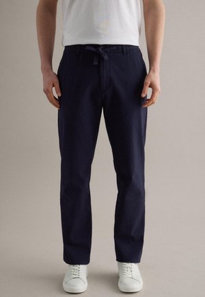 Hombre con pantalones azul marino texturizados con cintura de cordón, zapatillas blancas y una camisa blanca de manga corta, de pie contra un fondo liso.