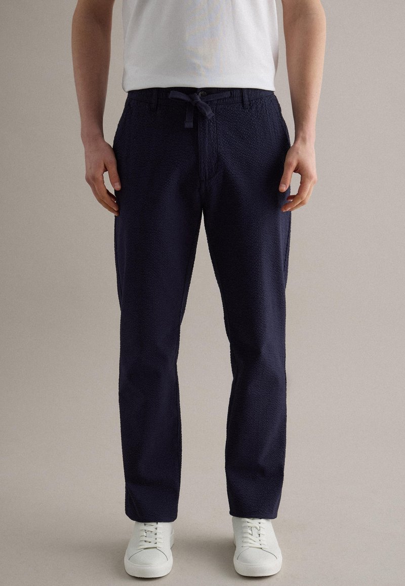 Man draagt getextureerde marineblauwe broek met trekkoord in de taille, witte sneakers en een wit T-shirt met korte mouwen, staand tegen een effen achtergrond.