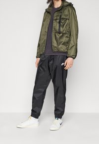 Olivgrön jacka med dragkedja och två framfickor, svarta träningsbyxor och vita sneakers med blå detaljer. Släta, lätta texturer.