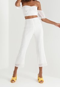 Biały dwuczęściowy strój składający się z krótkiego topu z odkrytymi ramionami i bufiastymi rękawami oraz zwiewnych spodni z szerokimi nogawkami. Żółte sandały na szpilkach.