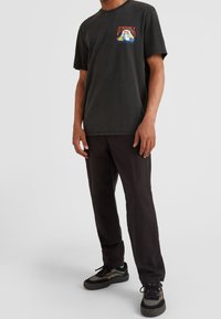 T-shirt nera a maniche corte con stampa grafica sul lato sinistro del petto, abbinata a pantaloni neri e sneakers multicolori.