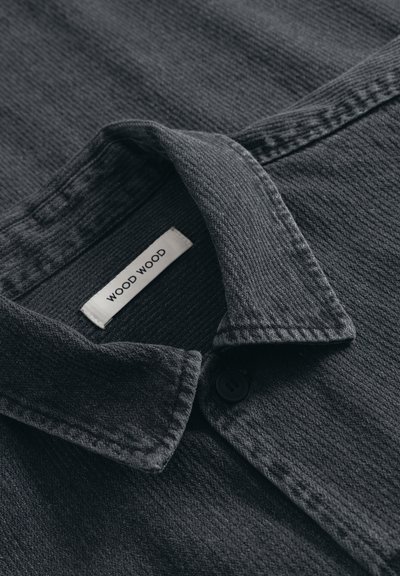 Crna pamučna košulja s teksturiranom površinom, šiljatim ovratnikom i kopčanjem na gumbe. Sadrži ušivenu etiketu na kojoj piše "WOOD WOOD".