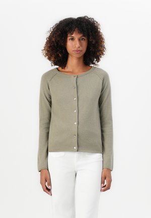 Frau mit lockigem Haar, die eine geknöpfte, olivgrüne Cardigan und weiße Hosen trägt, vor einem schlichten weißen Hintergrund steht.