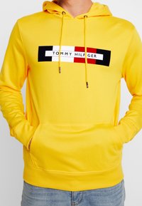 Sudadera amarilla brillante con bolsillo frontal y cordones, que presenta un logotipo rectangular de Tommy Hilfiger en negro, blanco y rojo en el pecho.