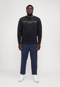 Black sweatshirt with "Tommy Hilfiger EST. 1985" logo, paired with navy pants and white sneakers, standing against a plain background.