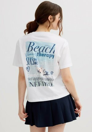 Mujer con cabello castaño recogido en una coleta, vistiendo una camiseta blanca con texto azul y gráfico de concha, combinada con una falda plisada azul marino.