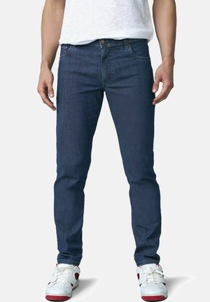 Mann trägt dunkelblaue Slim-Fit-Jeans, weiße Sneakers mit roten Akzenten und ein schlichtes weißes T-Shirt vor einfachem Hintergrund.