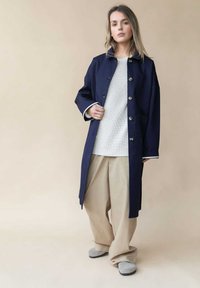 Marineblauer langer Mantel mit großen Knöpfen, getragen über einem weißen strukturierten Pullover und beigen weiten Hosen. Mit Kragen und zwei Seitentaschen.