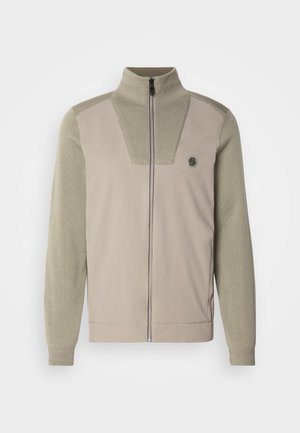 Šviesiai smėlio spalvos striukė su aukštu apykakle, užtrauktuku ir tekstūruotomis žaliomis rankovėmis. Yra krūtinės panelė ir logotipo detalė.