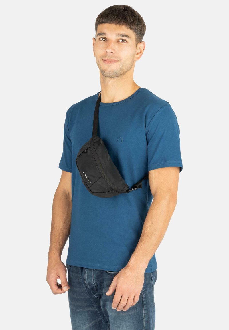 Schwarze Bauchtasche mit verstellbarem Riemen und einem Fach mit Reißverschluss. Getragen über einem blauen T-Shirt und kombiniert mit Jeans.
