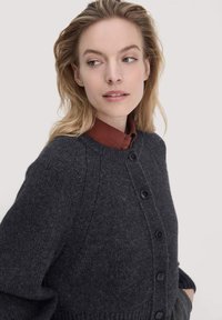 Frau, die seitlich schaut und eine dunkelgraue, geknöpfte Strickjacke über einem rostfarbigen Hemd mit Kragen trägt, vor einem hellen, einfarbigen Hintergrund.