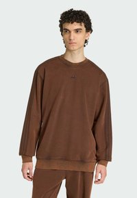 Hnedá mikina s okrúhlym výstrihom, m�äkká textura, rebrované manžety a vyšívané logo na hrudi. Obsahuje tri pruhy pozdĺž rukávov.