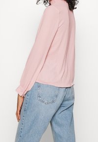 Chemise rose clair à manches longues avec un col montant, en tissu doux. Dotée de poignets boutonnés et d'un ourlet arrondi, portée avec un jean bleu clair.