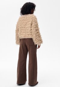 Osoba z kręconymi włosami odwrócona w stronę ściany, ubrana w beżowy, teksturowany sweter i szerokie brązowe spodnie na tle gładkiej białej ściany.