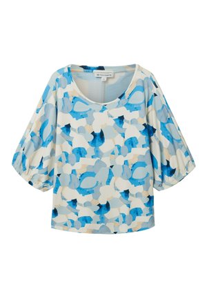 Kobieta w luźnym, wzorzystym niebiesko-beżowym topie, jasnoniebieskich szerokich dżinsach oraz brązowych sandałach na dwóch paskach, stoi z jedną ręką w kieszeni.