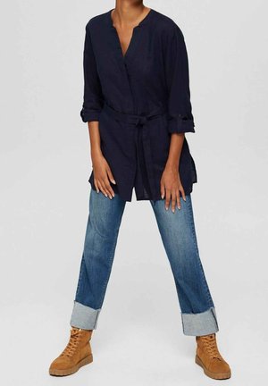 Granatowa tunika z paskiem wiązanym w talii, długimi rękawami i mankietami zapinanymi na guziki, zestawiona z niebieskimi, podwiniętymi jeansami oraz jasnobrązowymi botkami sznurowanymi.