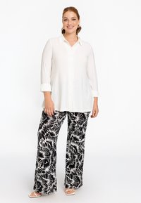 Witte lange mouwen knoopshirt gecombineerd met vloeiende zwart-witte bloemrijke broek; model draagt witte open hak sandalen.
