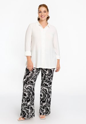 Weißes Langarmhemd mit Knöpfen, kombiniert mit fließenden Hosen mit schwarz-weißen Blumenmuster; Modell trägt weiße Sandalen mit offenen Zehen.