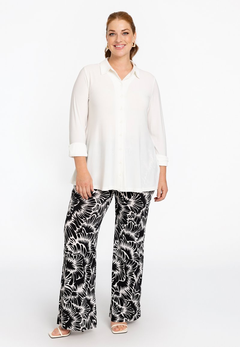 Witte lange mouwen knoopshirt gecombineerd met vloeiende zwart-witte bloemrijke broek; model draagt witte open hak sandalen.