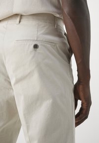 Beige bomullbyxor med en slät textur, med en bakficka och knappdetalj, som bärs på en arm som vilar lätt mot tyget.