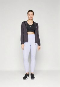 Young woman standing wearing black sports bra, lightweight black jacket, light purple leggings, and black Adidas sneakers on plain background.