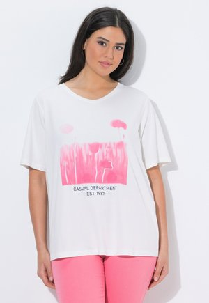 Frau mit dunklem Haar, die ein weißes T-Shirt mit pinkfarbenem Blumenmuster und dem Schriftzug "CASUAL DEPARTMENT EST. 1987" trägt, kombiniert mit pinken Hosen.