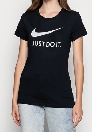 Kobieta nosząca czarną koszulkę Nike z białym logo Swoosh i napisem „JUST DO IT.”, w zestawieniu ze jasnoniebieskimi dżinsami.
