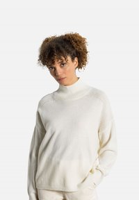 Maglione bianco ampio con collo alto, dettagli a coste e tessuto leggermente strutturato. Presenta spalle scese e maniche lunghe.
