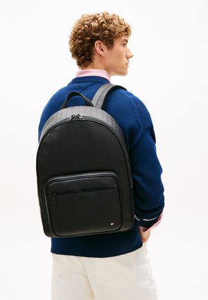 Junger Mann in blauem Pullover trägt einen strukturierten schwarzen Rucksack mit Vordertasche und kleinem Logo, seitlich auf weißem Hintergrund stehend.