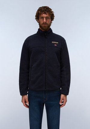 Mann mit lockigem Haar und Bart trägt eine dunkelblaue Fleecejacke mit Reißverschluss vorn und blaue Jeans vor einem schlichten, hellen Hintergrund.